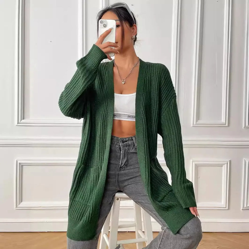 A woman wearing a dark green, loose-fit, mid-length acrylic cardigan with long sleeves and two front pockets.