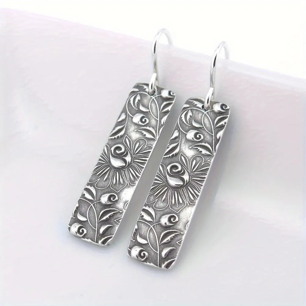 Silver geometric long drop earrings with a carved vine pattern on a white background.