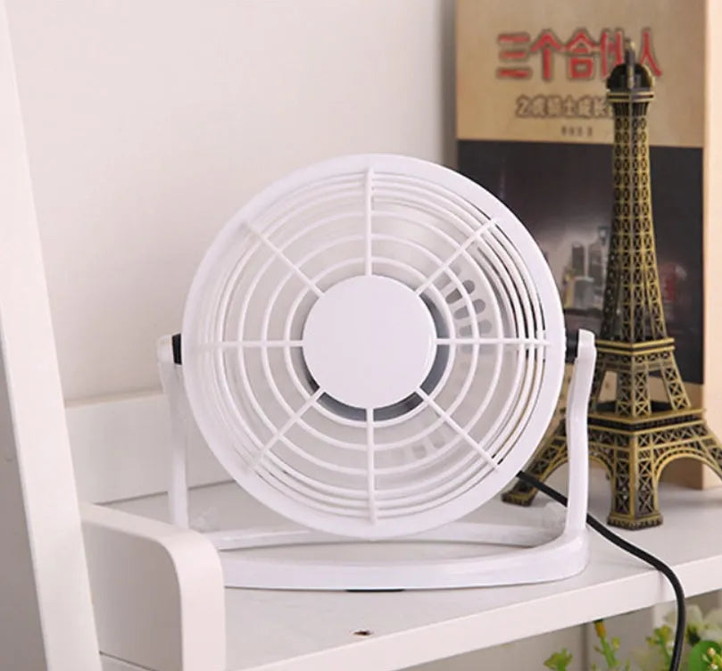 Mini black USB desk fan with adjustable tilt angle on a wooden desk next to a laptop.