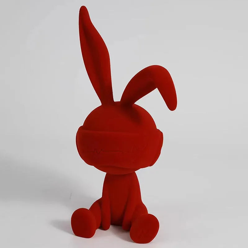 Green resin table ornament of a rabbit wearing a suede blindfold on a wooden shelf.