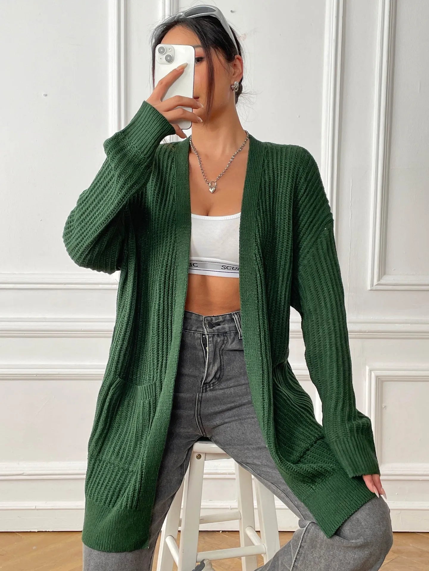 A woman wearing a dark green, loose-fit, mid-length acrylic cardigan with long sleeves and two front pockets.
