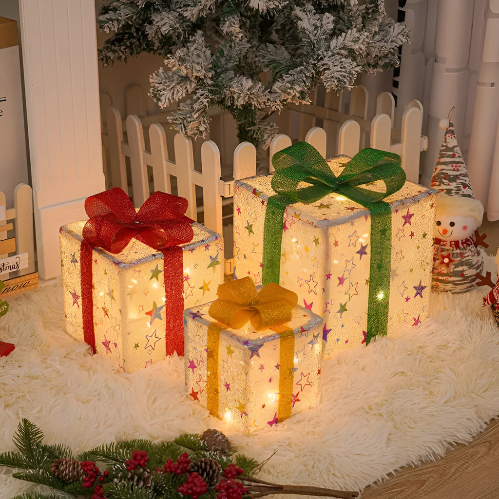 Set of three glittering pre-lit Christmas gift boxes with red bows, illuminated with warm white LED lights outdoors.