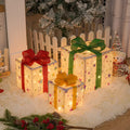 Set of three glittering pre-lit Christmas gift boxes with red bows, illuminated with warm white LED lights outdoors.