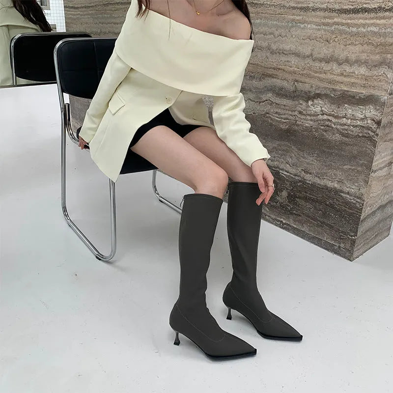 Black women's pointed toe below-the-knee boots with a stiletto heel and back zipper on a plain background.