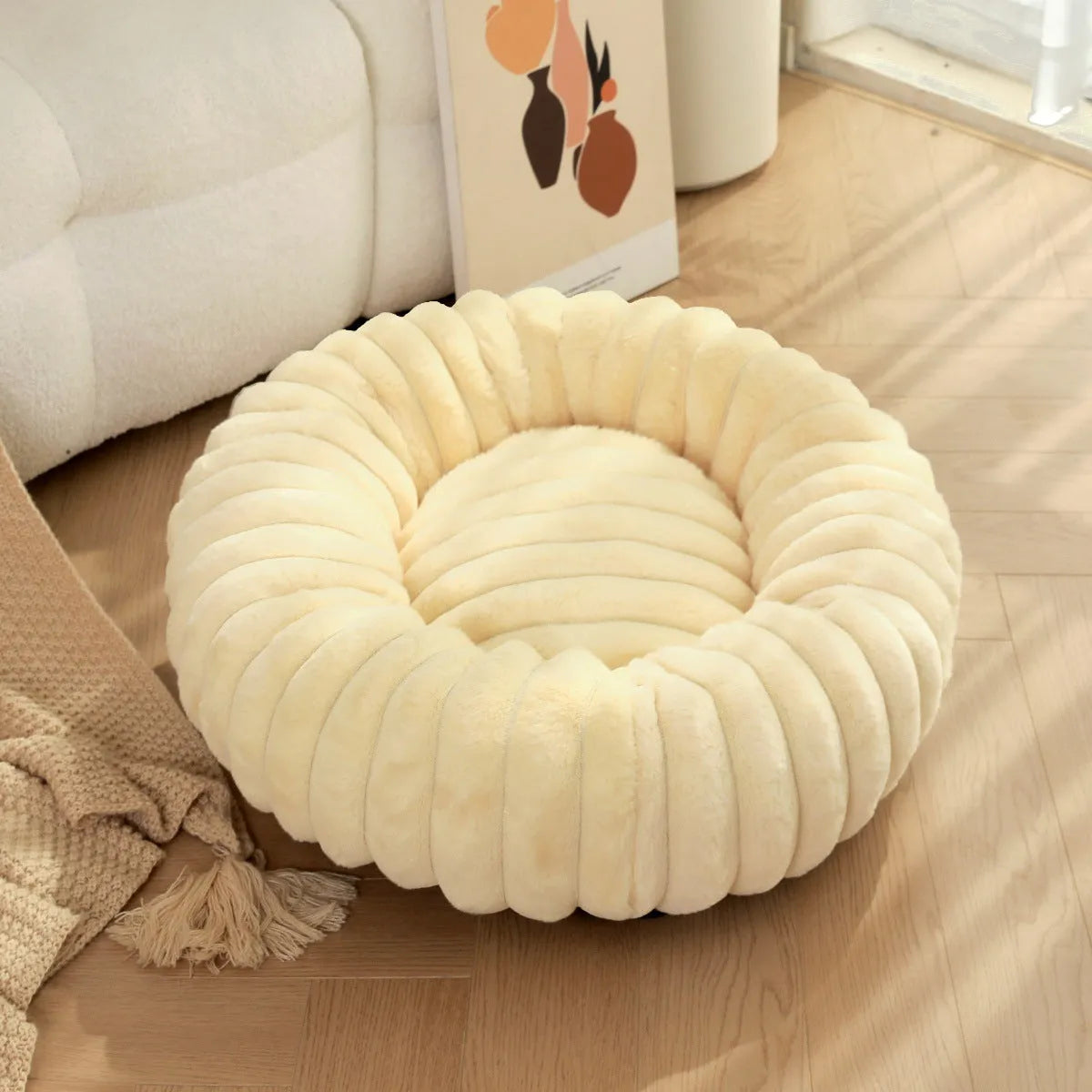 Round beige plush pet bed for a dog, shown in a medium size on a wooden floor.