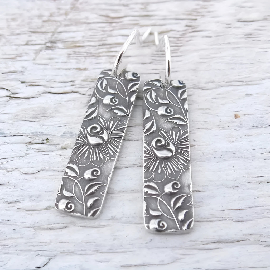 Silver geometric long drop earrings with a carved vine pattern on a white background.