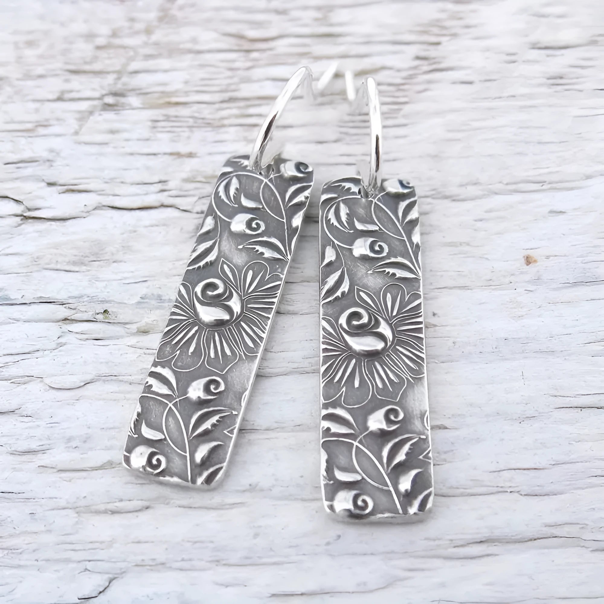 Silver geometric long drop earrings with a carved vine pattern on a white background.