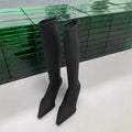 Black women's pointed toe below-the-knee boots with a stiletto heel and back zipper on a plain background.