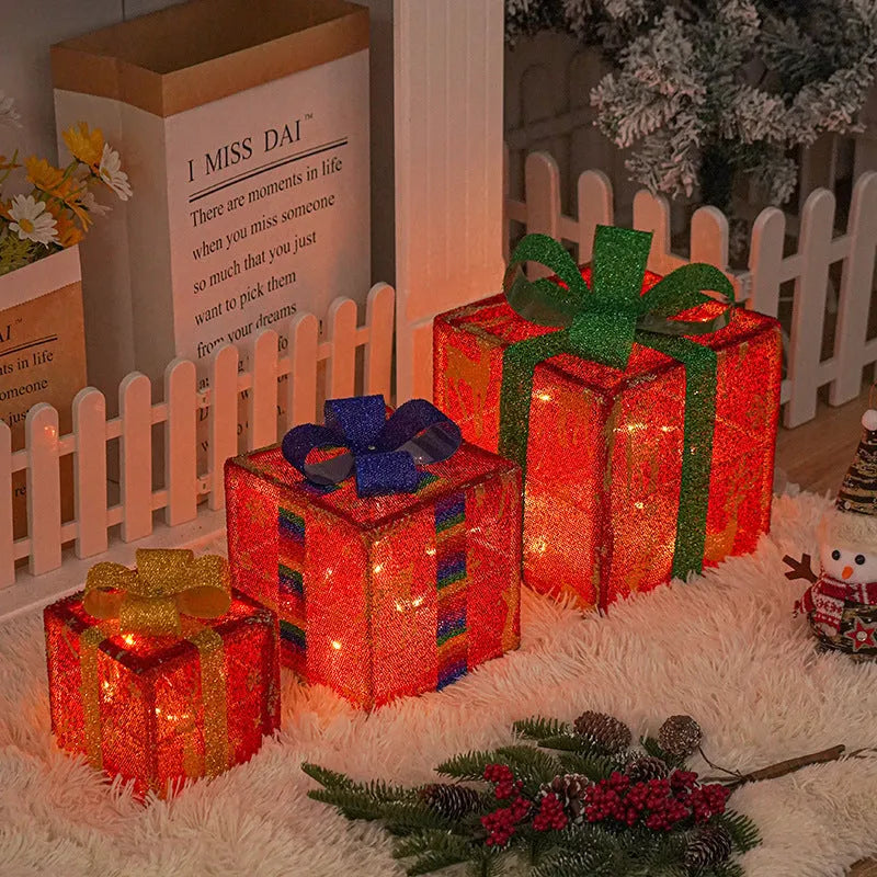 Set of three glittering pre-lit Christmas gift boxes with red bows, illuminated with warm white LED lights outdoors.