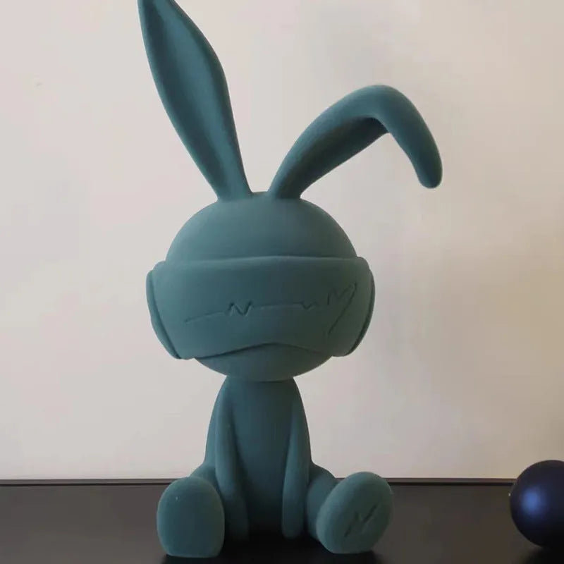 Green resin table ornament of a rabbit wearing a suede blindfold on a wooden shelf.