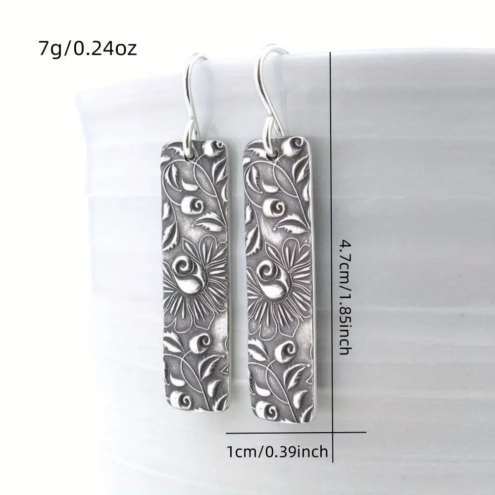 Silver geometric long drop earrings with a carved vine pattern on a white background.