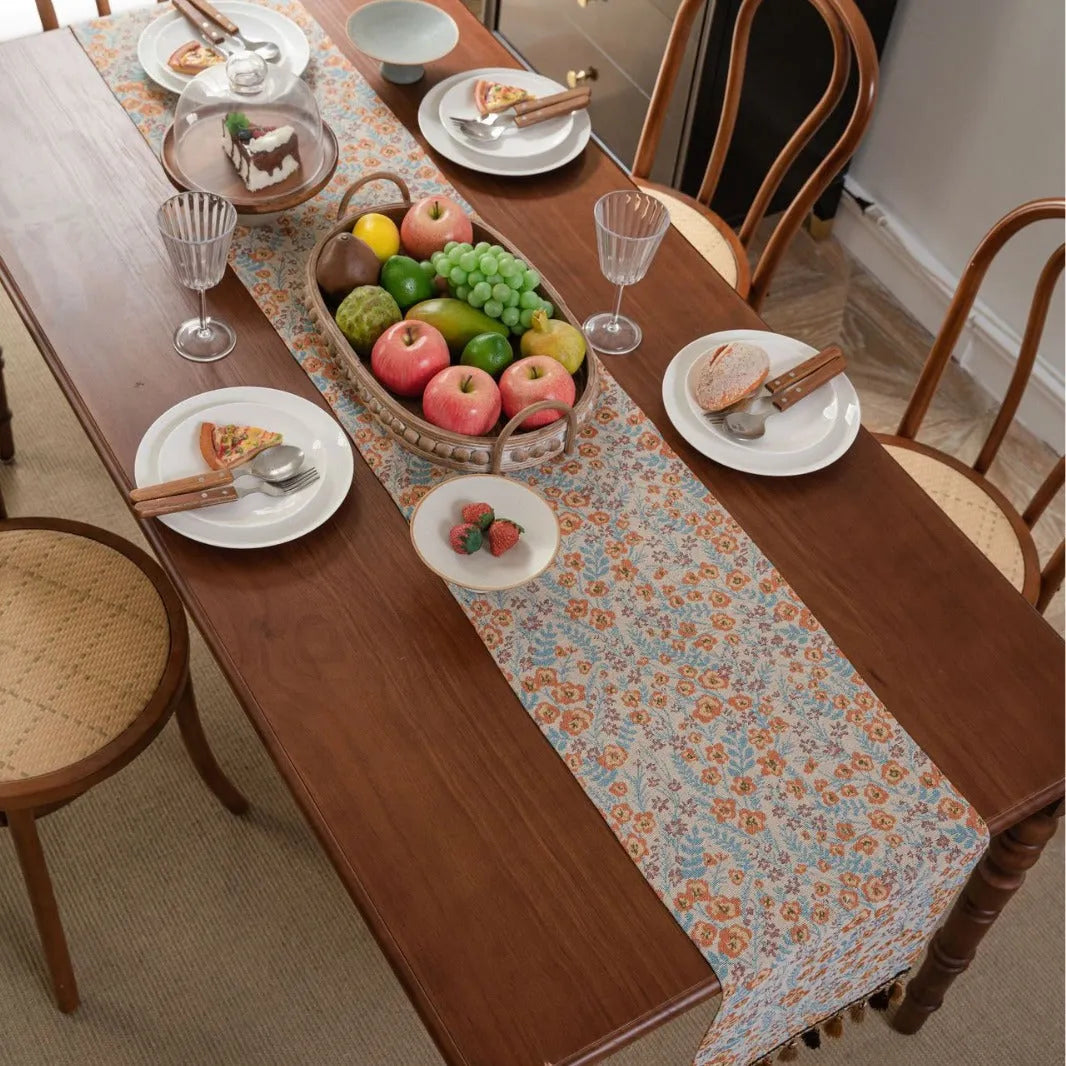Beige jacquard table runner with floral leaf pattern and long tassels on a dining table.