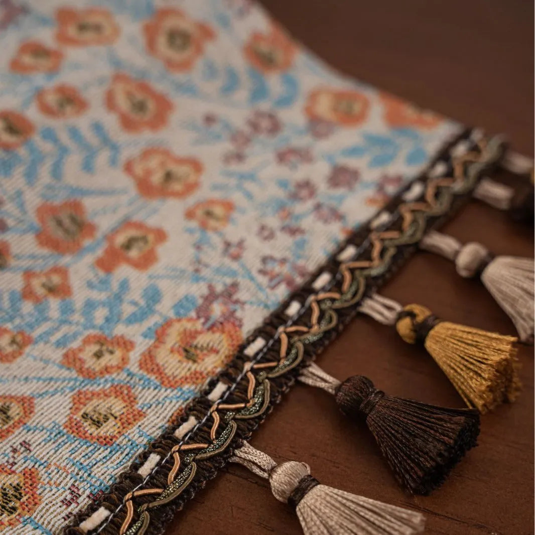 Beige jacquard table runner with floral leaf pattern and long tassels on a dining table.