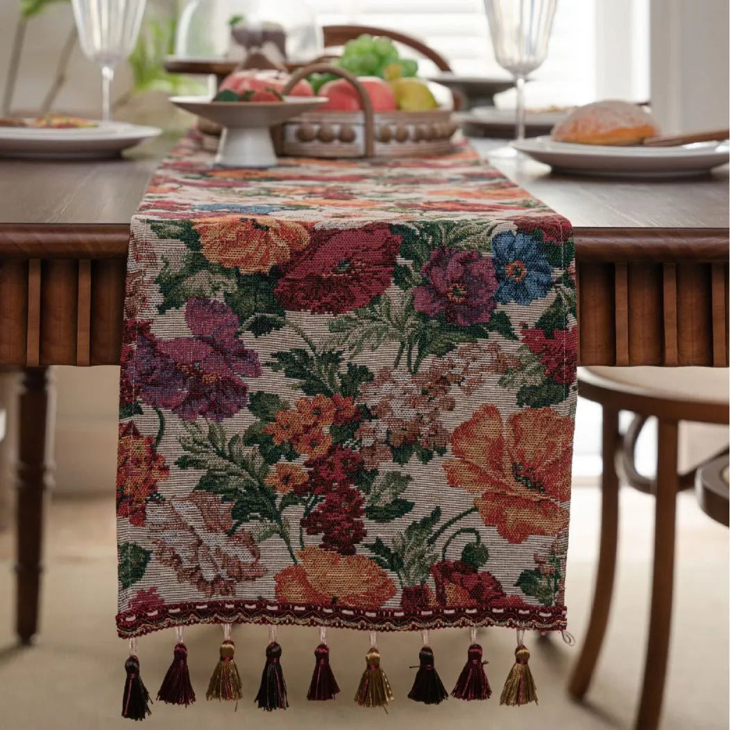 Beige jacquard table runner with floral leaf pattern and long tassels on a dining table.