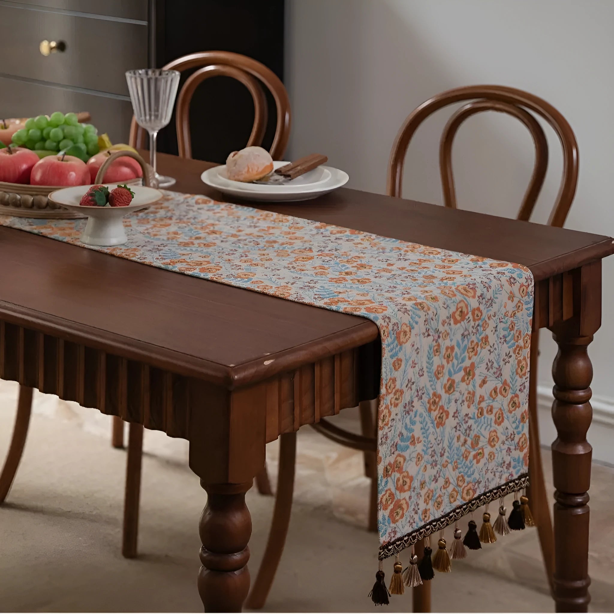 Beige jacquard table runner with floral leaf pattern and long tassels on a dining table.