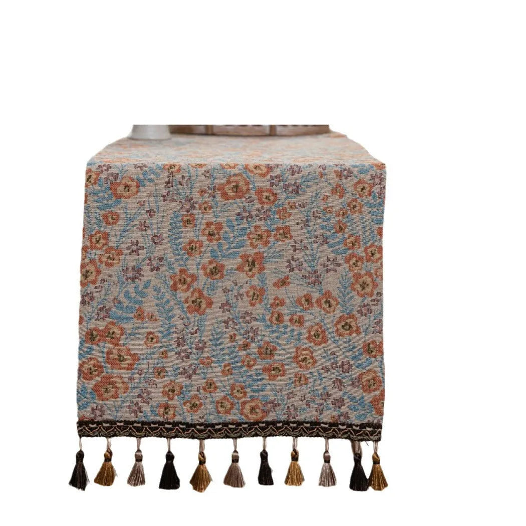 Beige jacquard table runner with floral leaf pattern and long tassels on a dining table.