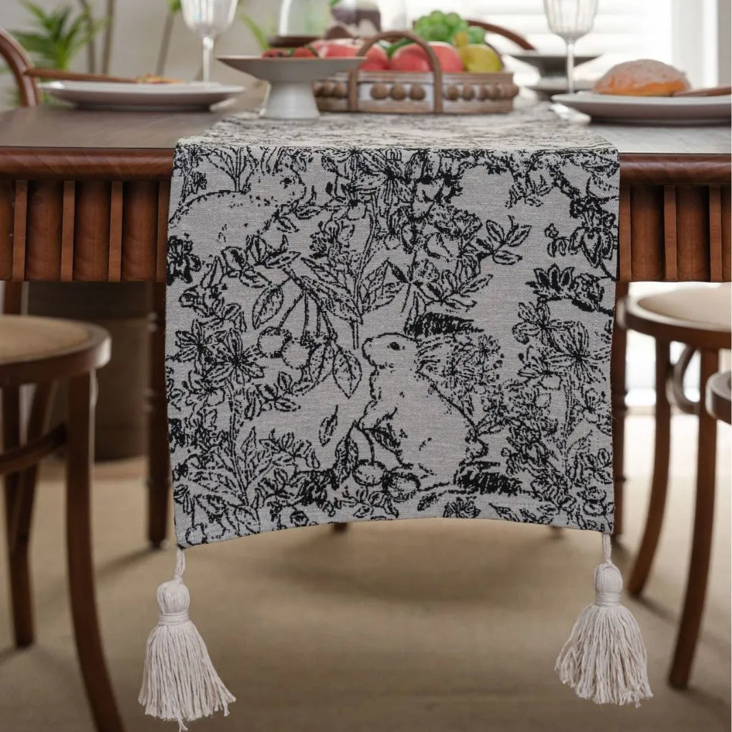 Beige jacquard table runner with floral leaf pattern and long tassels on a dining table.