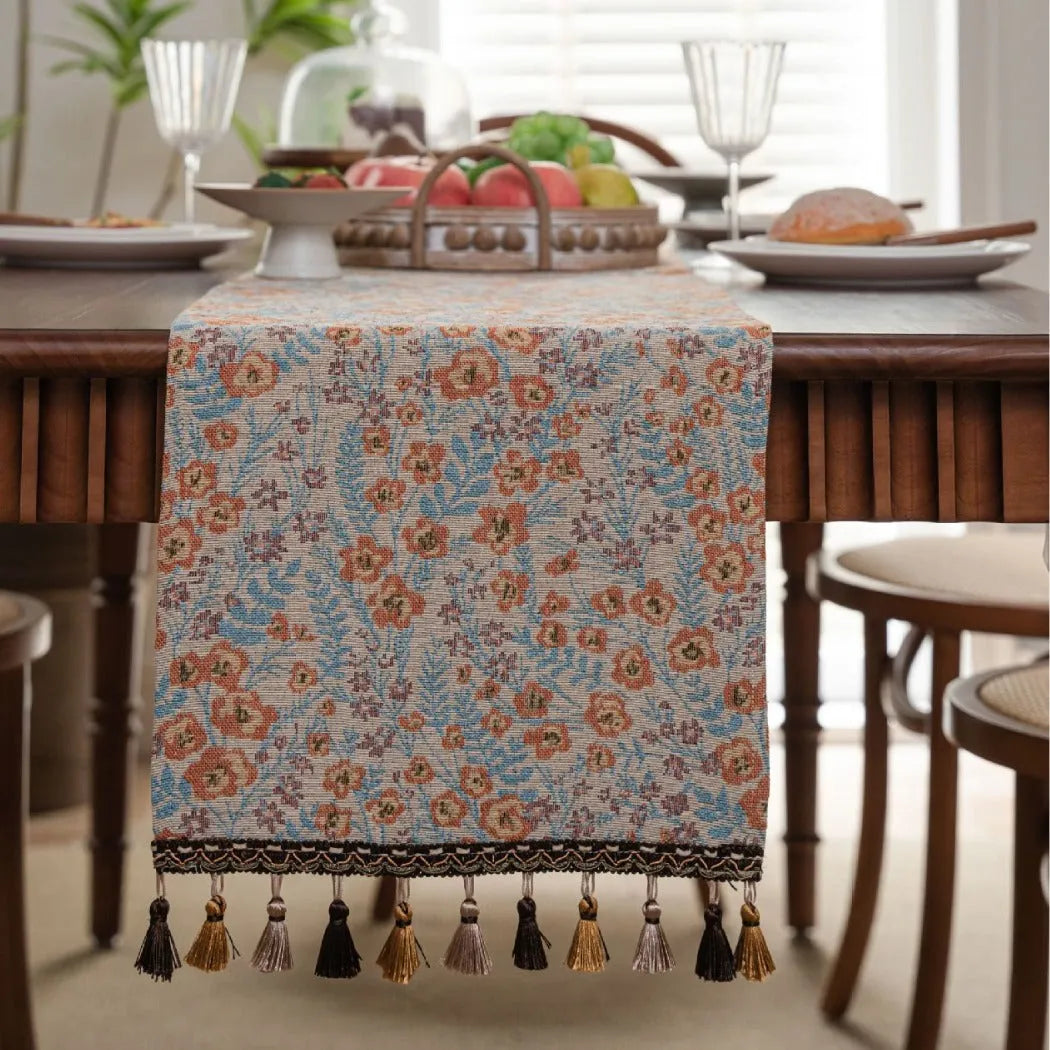 Beige jacquard table runner with floral leaf pattern and long tassels on a dining table.