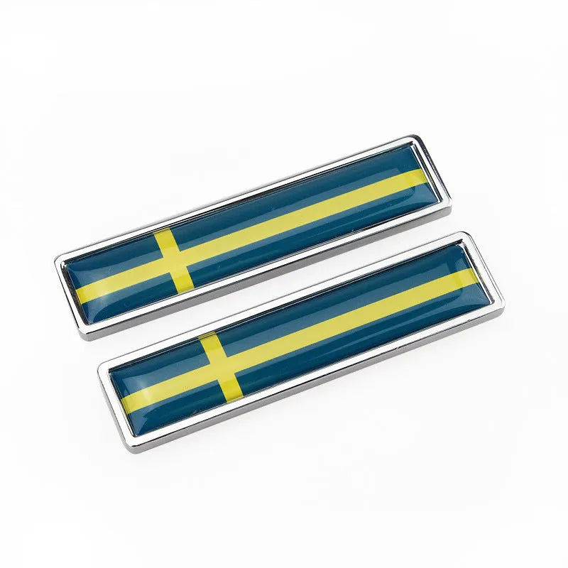 A pair of silver zinc alloy car badge labels with coloured national flag designs, shown at 58mm by 14mm scale.