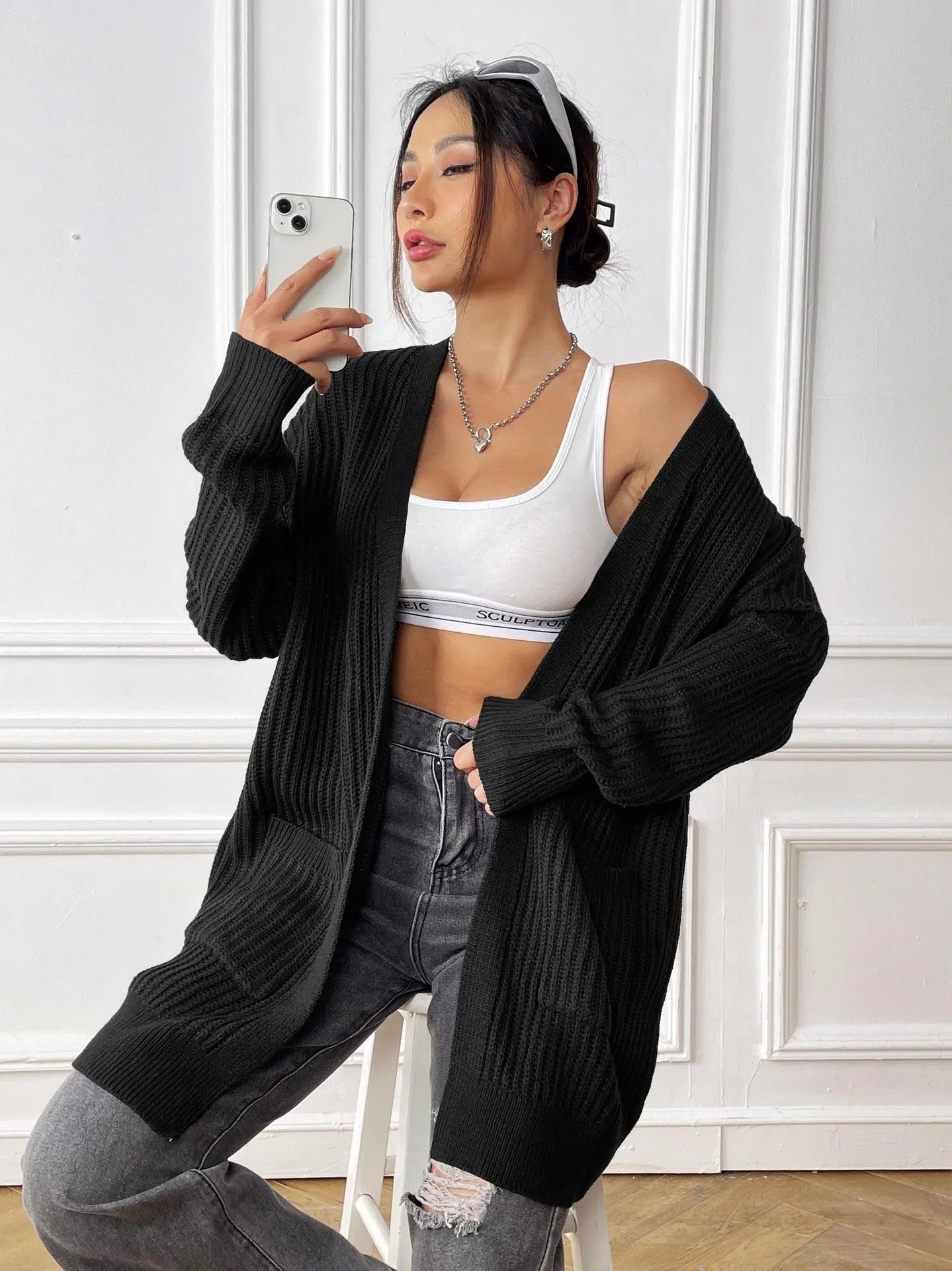 A woman wearing a dark green, loose-fit, mid-length acrylic cardigan with long sleeves and two front pockets.