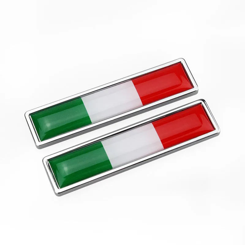 A pair of silver zinc alloy car badge labels with coloured national flag designs, shown at 58mm by 14mm scale.