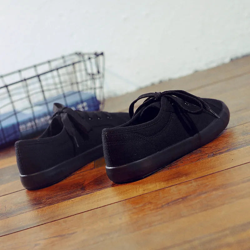 All black low-top canvas flat shoes with white laces and a rubber sole.