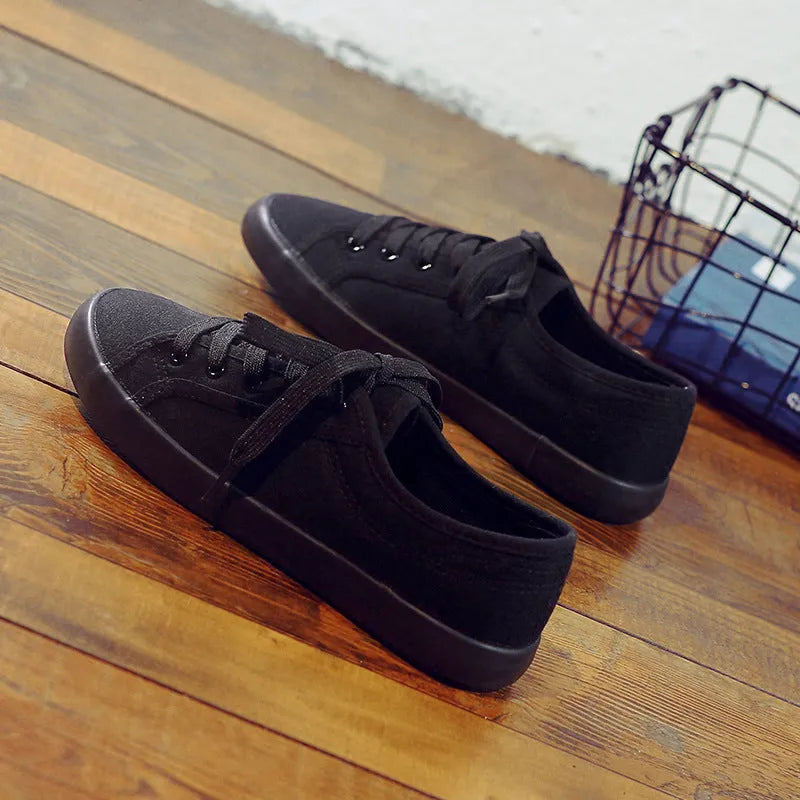 All black low-top canvas flat shoes with white laces and a rubber sole.