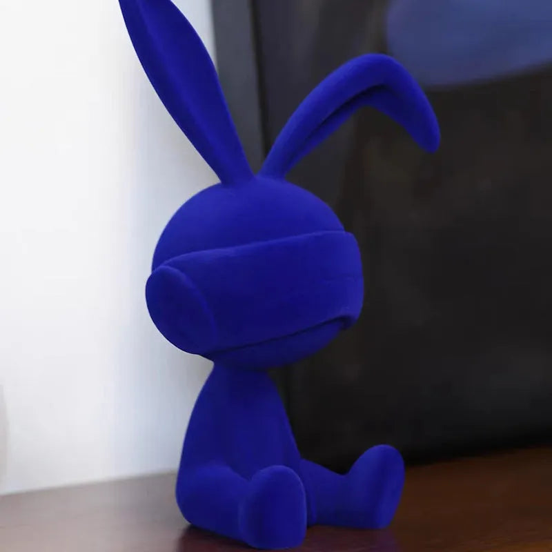 Green resin table ornament of a rabbit wearing a suede blindfold on a wooden shelf.