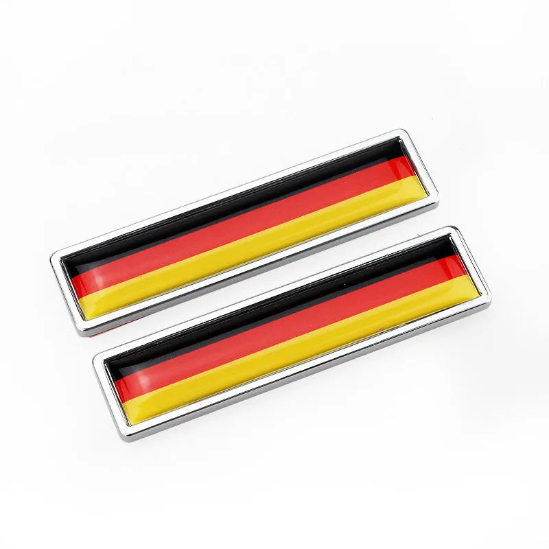 A pair of silver zinc alloy car badge labels with coloured national flag designs, shown at 58mm by 14mm scale.