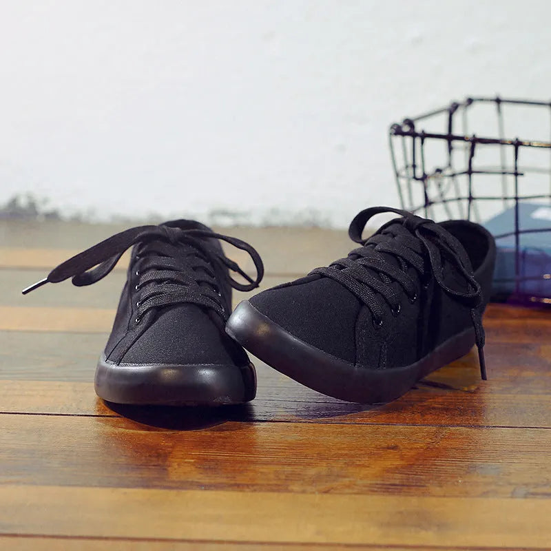 All black low-top canvas flat shoes with white laces and a rubber sole.